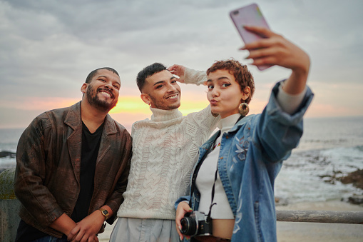 Smiling group of diverse young friends taking a selfie together on a causeway overlooking the ocean at sunset