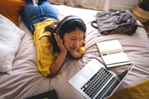 istock Asian college student at home lying on bed using laptop attending online university classes listening with headphones. 1492858045