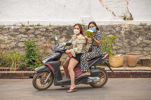 Luang Prabang, Laos - March 12th 2023: Two women on a motorbike driving down the main street and making fun with the photographer