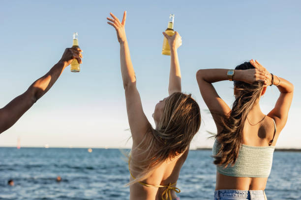 若い女性は海を見ながらビールを飲む ボトルを上げるパーティーは幸せ - party beach indian ethnicity adult ストックフ�ォトと画像
