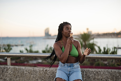 pretty african woman traveler, on the beach, eating an ice cream, wears a summer bikini.