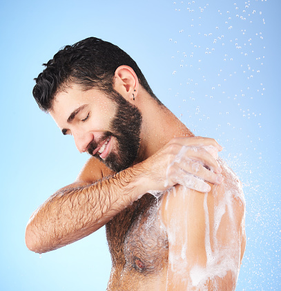 Shower, cleaning and man with water, smile and soap in studio for wellness, hygiene and grooming. Skincare, healthy skin and happy male with foam, bath cosmetics and washing body on blue background