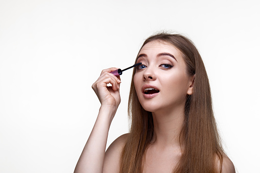 Beautiful young woman applying black mascara on long thick eyelashes with a brush and making night makeup on white background