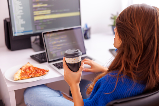 Female programmer sitting at her desk in home office, taking a break while working remotely from home drinking coffee and eating pizza