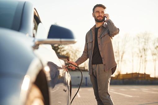 Business call by phone. Man is standing near his electric car outdoors.