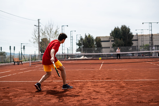 men tennis player with orange short is shooting backhand on clay court focus on foreground horizontal tennis still