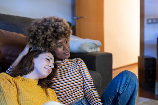 Happy multiracial lesbian couple watching tv at home hugging. Two relaxed females sitting on the floor watching a movie.