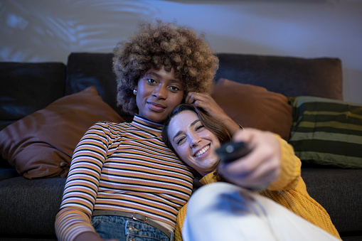 Multiracial lesbian women looking and pointing to camera with the remote control. Lovely couple relaxing on the floor of a living room smiling and hugging while one of them is changing tv channel. Focus on girls.