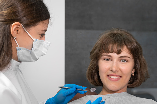 Young smiling woman is sitting in a dental chair and looking into the camera, the dentist is holding dental instruments for examining the oral cavity