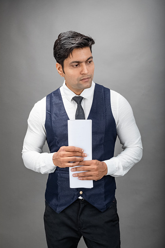 Indian young business man giving presentation on a topic holding folder in hand in formal wear wearing white shirt, black tie, black coat against grey background. Male model.