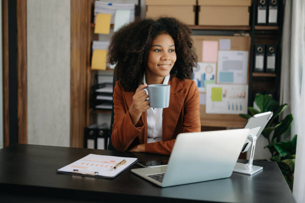 istock Confident beautiful African businesswoman typing laptop computer and digital tablet while holding coffee at office 1492884959