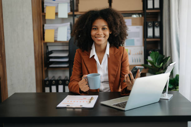 istock Confident beautiful African businesswoman typing laptop computer and digital tablet while holding coffee at office 1492884979