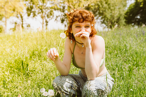 red-haired teenage girl, in swimsuit and shorts, sits on grass, blows on dandelion, winces from an allergy to fluff, on sunny summer day