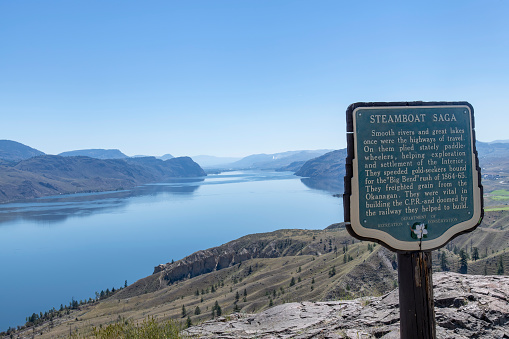 Kamloops Lake, BC, Canada-August 2022; Panoramic view over Kamloops Lake  and surrounding mountain on Thompson River, west of Kamloops with in front plaque for Steamboat Saga against a clear blue sky