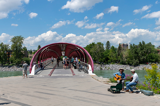 Calgary, AB, Canada-August 2022; Entrance view red and white colored helical shaped Peace Bridge across Bow River by design of architect Santiago Calatrava with two musicians playing guitar in front