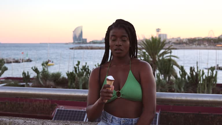 pretty african woman traveler, on the beach, eating an ice cream, wears a summer bikini.