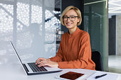 Portrait of happy and successful female programmer inside office at workplace, worker smiling and looking at camera with laptop, blonde businesswoman is satisfied with results of achievements at work