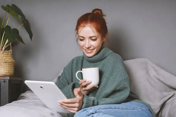 woman in turleneck sweater relaxing at home using tablet computer for entertainment or communication and drinking coffee