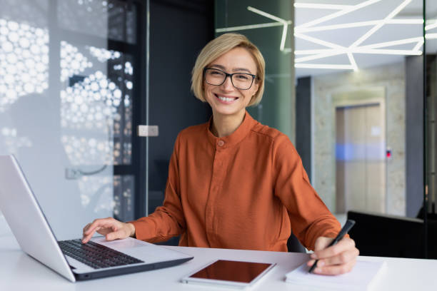 istock Successful and happy business woman looking at camera sitting inside office working with documents, portrait of satisfied woman writing in notebook and working using laptop at work 1492896574