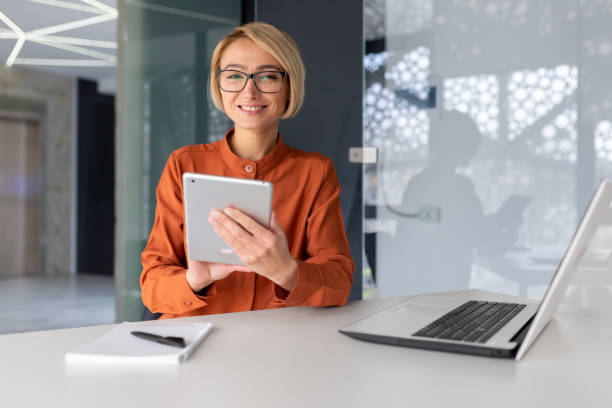 portrait d’une jeune étudiante assise dans un bureau, un espace de coworking, une bibliothèque à une table avec un ordinateur portable et utilisant une tablette pour étudier. souriante, elle regarde la caméra. - looking at camera smiling desk isolated photos et images de collection