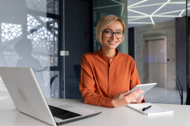 porträt einer jungen frau, die im büro an einem tisch mit einem laptop sitzt und ein tablet benutzt. er schaut in die kamera, lächelt. - glasses touchpad professional occupation women stock-fotos und bilder