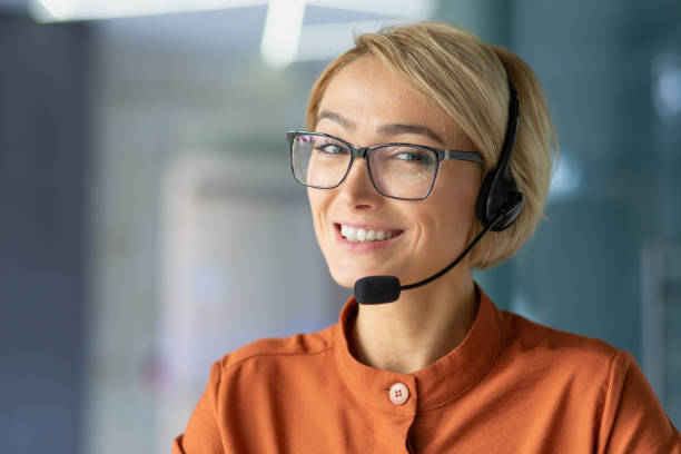 close-up portrait of young beautiful blonde woman at workplace, call center worker smiling and looking at camera, operator using headset phone for video call and online customer consultation support. - hotel reception customer service representative headset receptionist imagens e fotografias de stock