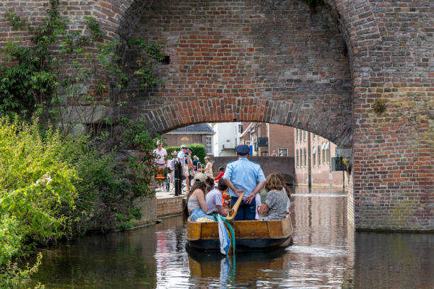 Zutphen, the Netherlands-August 2022; Close up of a small cruise boat with captain standing going under the medieval Berkel Poort or water gate and fortified wall of the city with tourists on board