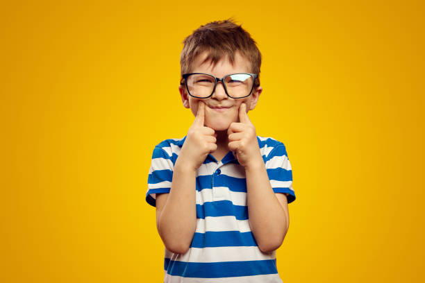 istock Kid wearing glasses and blue striped polo, touching cheeks with both index finger against yellow 1492902274