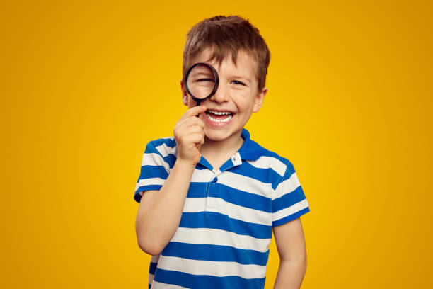 istock Cute kid in casual wear looking at camera through magnifier while smiling against yellow background 1492903251