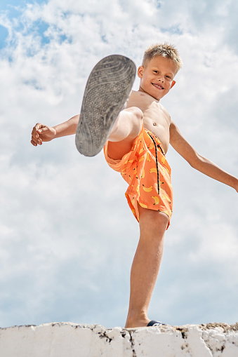 Junior schoolboy with delighted and smiling expression on high pier against blue sky with clouds. Boy raises foot upward enjoying spending summer holidays at seaside
