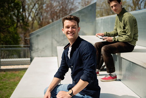 Portrait of college student relaxing on rooftop outdoors in springtime. Focus on white young man with smile, dressed in casual clothes. Another male student working in the background. Horizontal waist up outdoors shot with copy space. This was taken in Quebec, Canada.