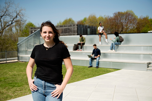 Portrait of college student on a rooftop outdoors in springtime. White young woman with long hair, dressed in casual clothes. Small group of students sitting in stairs in the background. Horizontal waist up outdoors shot with copy space. This was taken in Quebec, Canada.