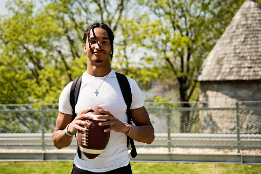 Portrait of college student holding a football outdoors in springtime. Black young man with braids, dressed in casual clothes with a backpack. Horizontal waist up outdoors shot with copy space. This was taken in Quebec, Canada.
