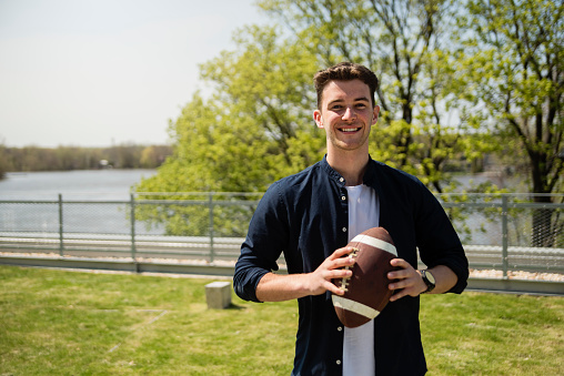Portrait of college student holding a football outdoors in springtime. White young man with smile, dressed in casual clothes. Horizontal waist up outdoors shot with copy space. This was taken in Quebec, Canada.