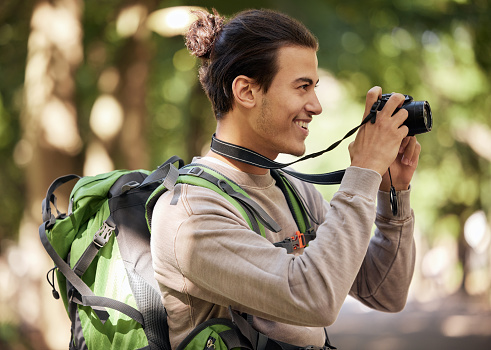 Photographer man, camera and outdoor park with smile, focus or adventure with creative vision. Young nature journalist, photography expert and bird watching with tech, hiking or backpack for research