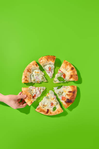 istock Four cheese pizza with tomatoes and spinach, cut into 6 slices on a bright green background. A woman's hand grabs a slice. Top view, minimalist style. Great for modern food photography, takeout. 1492929001