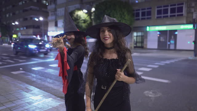Two Colombian Girls In Witch Halloween Costumes With Broom Sticks Looking At Camera Smiling While Standing At The City Sidewalk At Night. -  Medium Closeup Shot