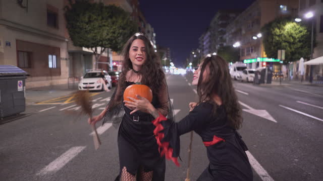 Two Lovely Colombian Girls In Witch Costume With Broomsticks, One Holding A Pumpkin Having Fun Walking Down The Street Road At Night, Valencia, Spain. - Full Shot