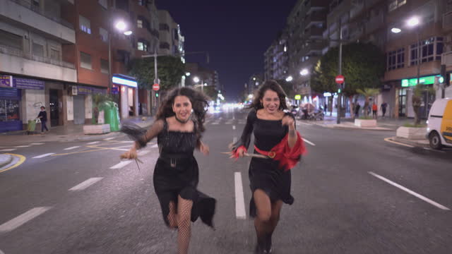 Two Beautiful Colombian Women In Witch Costumes With Broomsticks Walking Fast And Start Running Towards The Camera In City Street At Night On Halloween Holiday In Valencia, Spain - Pull-Back Shot