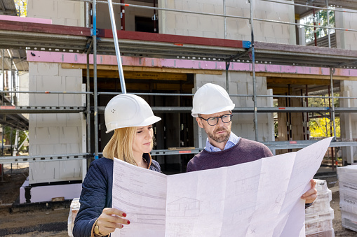 Mid adult couple with hardhats standing outside their new home under construction examining the blueprint. Homeowners standing outside their new under construction house and inspecting progress.