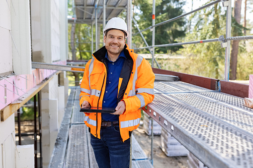 Portrait of a happy male engineer standing at construction site. Architect in  hardhat and reflective clothing holding digital tablet outside under construction building site.