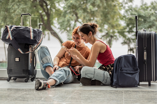 Young friends woman using phone sitting on ground while waiting at airport