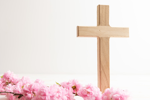 Small wood Christian cross with a branch with green leaves and pink flower blossoms on a white background with copy space