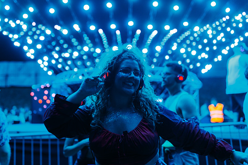 A young girl is dancing at the dj set at a festival, wearing casual clothes and listening to the music on a set of headphones.