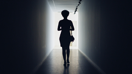 Rear view of a silhouetted young businesswoman walking down a dark office corridor to a lit doorway