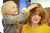 Cute little boy making funny hairstyle for his young mother. Preschooler child brush hair of his mom and play as if he works in a beauty salon.