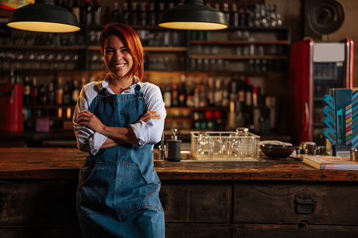 Portrait of a Hispanic female business owner proudly standing in front of the bar with arms crossed and smiling.