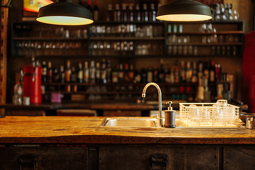 Indoor warm photography of a bar counter with the sink and blurred shelves with liquor bottles in the background.