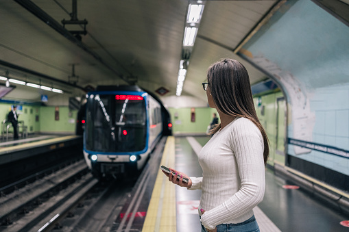 Young brunette woman watching from the platform the arrival of the subway while holding her mobile phone.