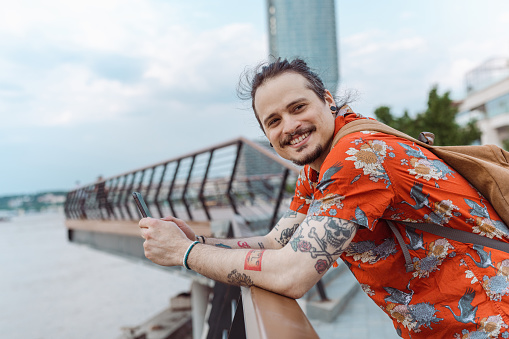 A cheerful tourist in the shirt is feeling happy. He is standing in the city center with his arms on the fence and smiling.